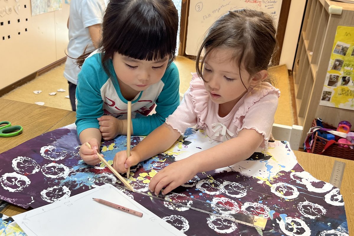 共同制作した絵の上で、定規と鉛筆を使って形を写し取る子どもたち