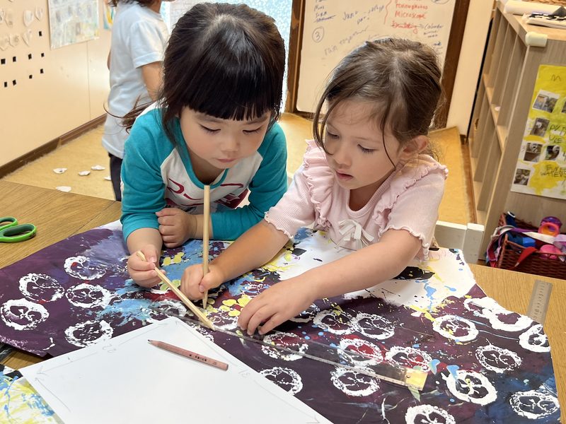 共同制作した絵の上で、定規と鉛筆を使って形を写し取る子どもたち